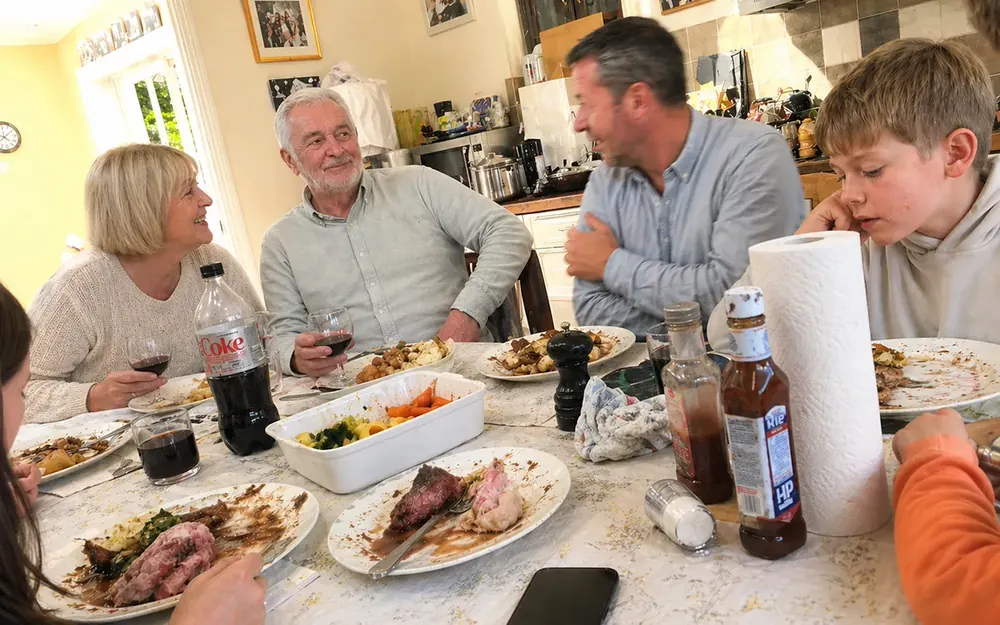 A British family around a roast dinner table mid-meal. The grandfather smiles faintly while the conversation moves on without him.